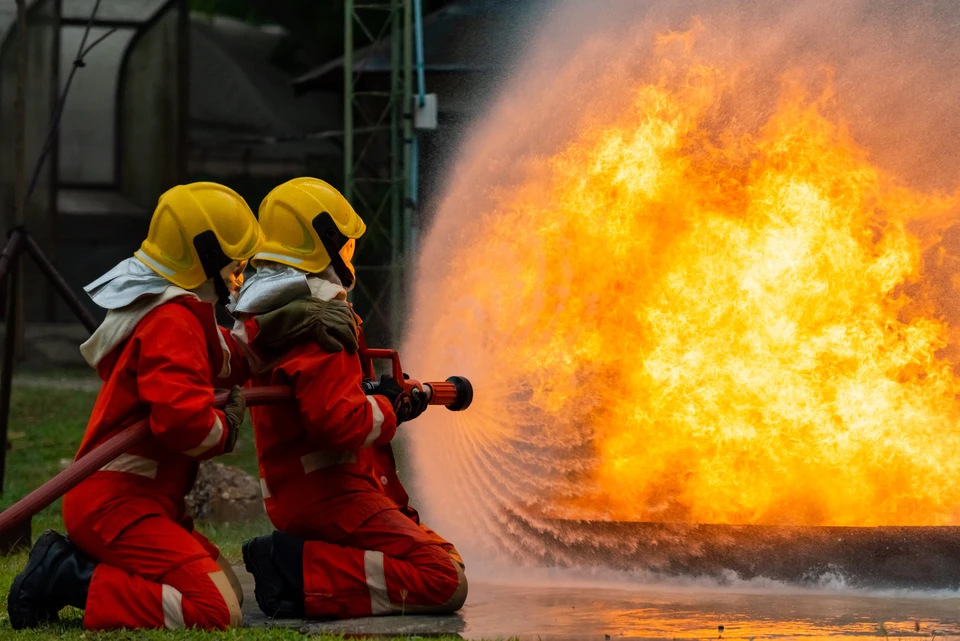 Deux pompiers en combinaisons de protection rouge et casques jaunes combattent un incendie industriel avec une lance à incendie, projetant de l'eau sur d'importantes flammes oranges dans un environnement extérieur